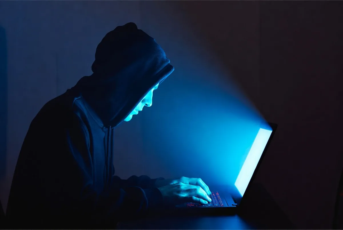 A hooded figure at a laptop in a dark room