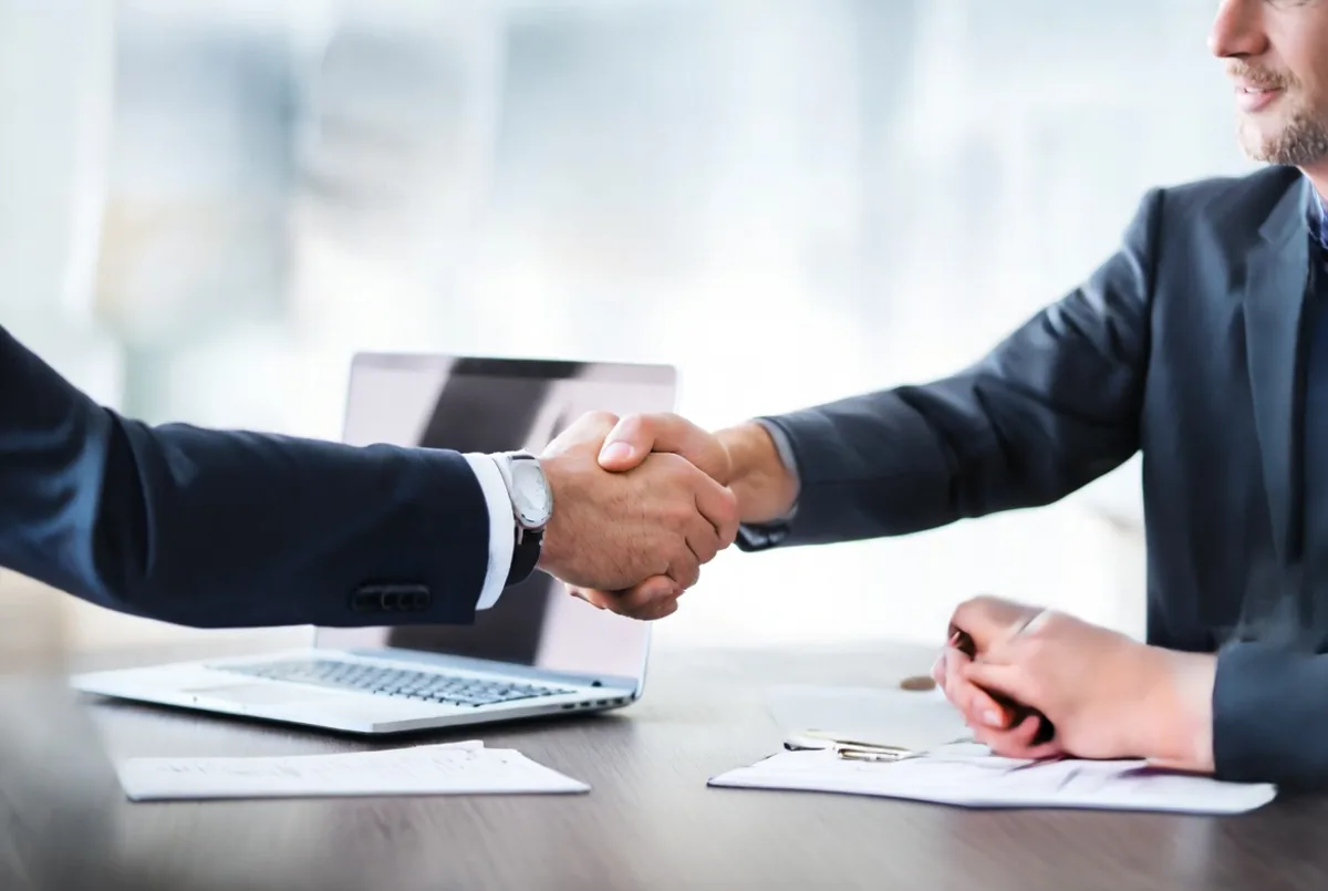 A handshake across a desk with a laptop nearby