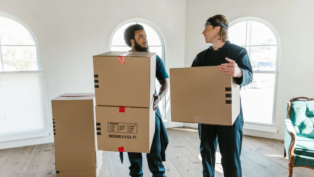 Two professional movers carrying cardboard boxes through a bright room with large arched windows